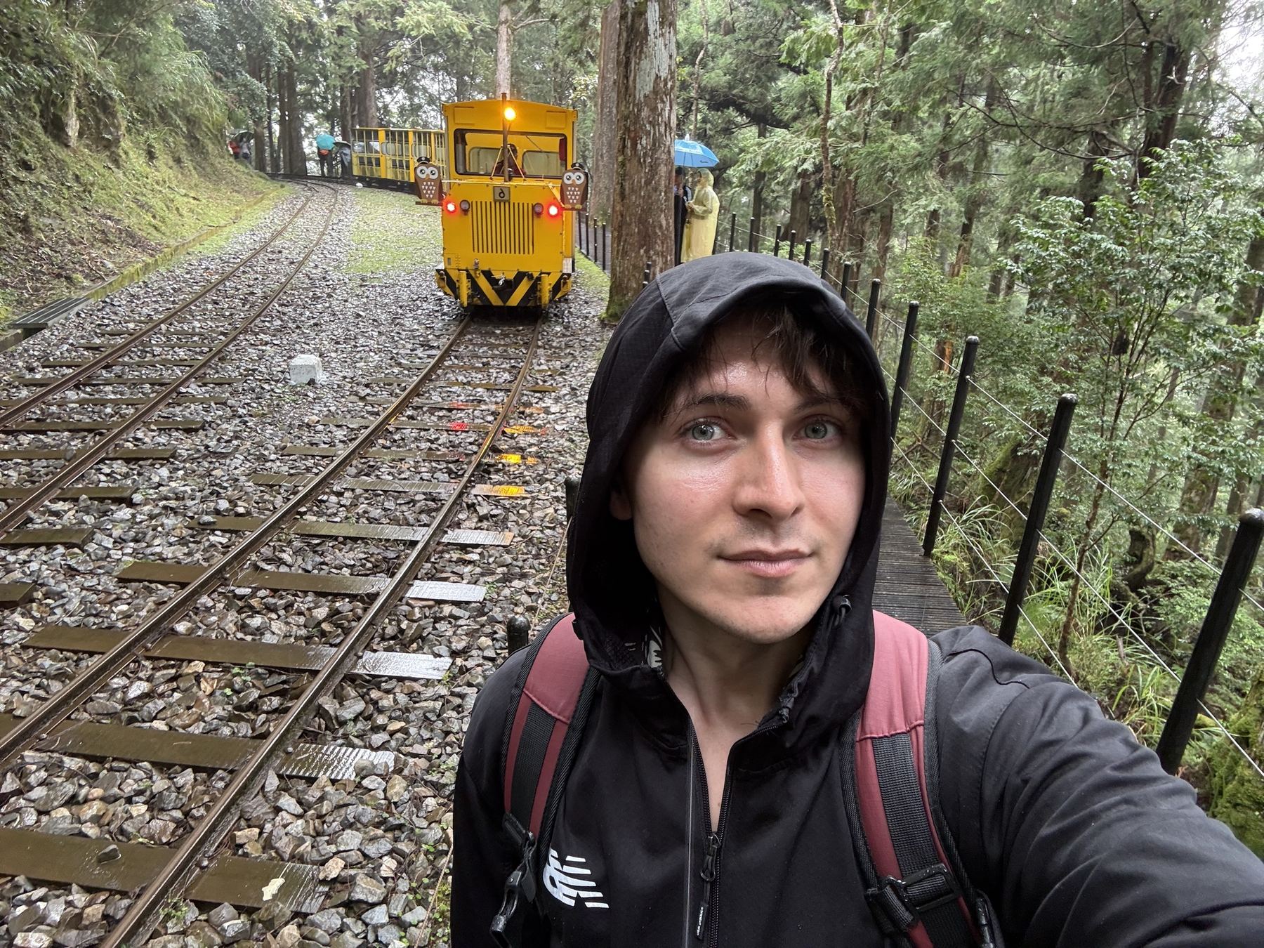 Maciej in a rain hood beside a yellow forest train in Taiwan